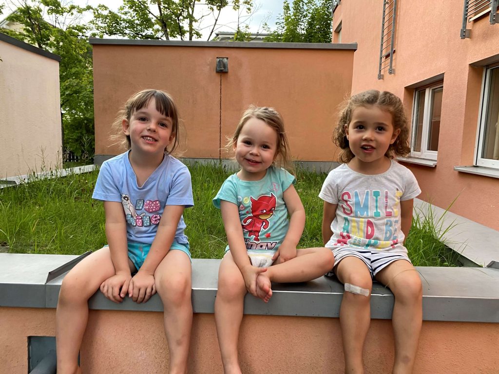 Drei kleine Kinder sitzen nebeneinander auf einer niedrigen Mauer im Garten und lächeln in die Kamera.